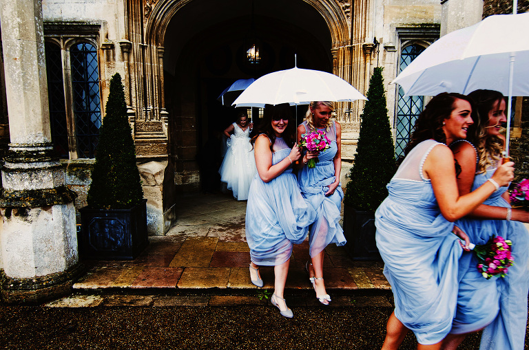 Bride and bridesmaids with white wedding umbrellas outside the entrance of hengrave hall on the way to the church.